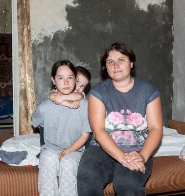 In einer kargen Wohnung sitzen eine Frau und zwei Kinder auf der Kante des Bettes.