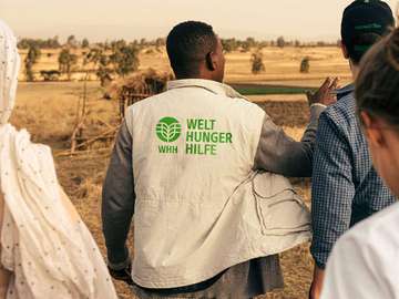 Ein Mitarbeiter der Welthungerhilfe zeigt in die Landschaft. Er ist von hinten zu sehen und trägt eine Weste mit dem neuen Logo der Welthungerhilfe.