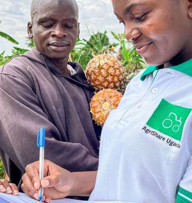 Titelfoto des Jahresbericht 2021. Eine Welthungerhilfe-Mitarbeiterin mit AgriShare-T-Shirt macht Notizen, während ein Mann im Hintergrund zuschaut. Er hält zwei Ananas in den Händen.