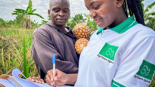 Titelfoto des Jahresbericht 2021. Eine Welthungerhilfe-Mitarbeiterin mit AgriShare-T-Shirt macht Notizen, während ein Mann im Hintergrund zuschaut. Er hält zwei Ananas in den Händen.