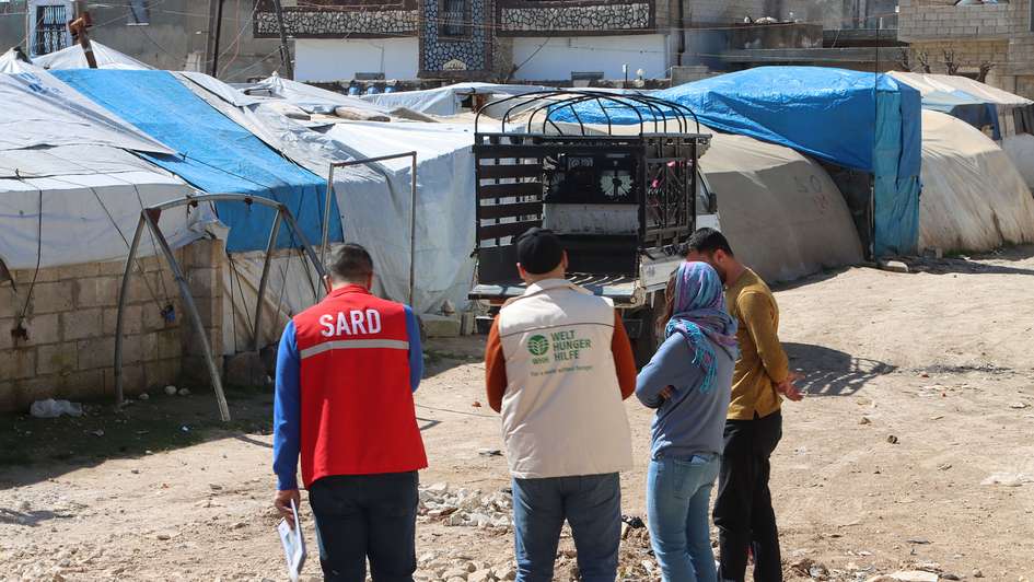 Four people, two of them in the uniforms of aid organizations, in a refugee camp in Syria. The surroundings consist of makeshift shelters covered with blue and white tarpaulins.